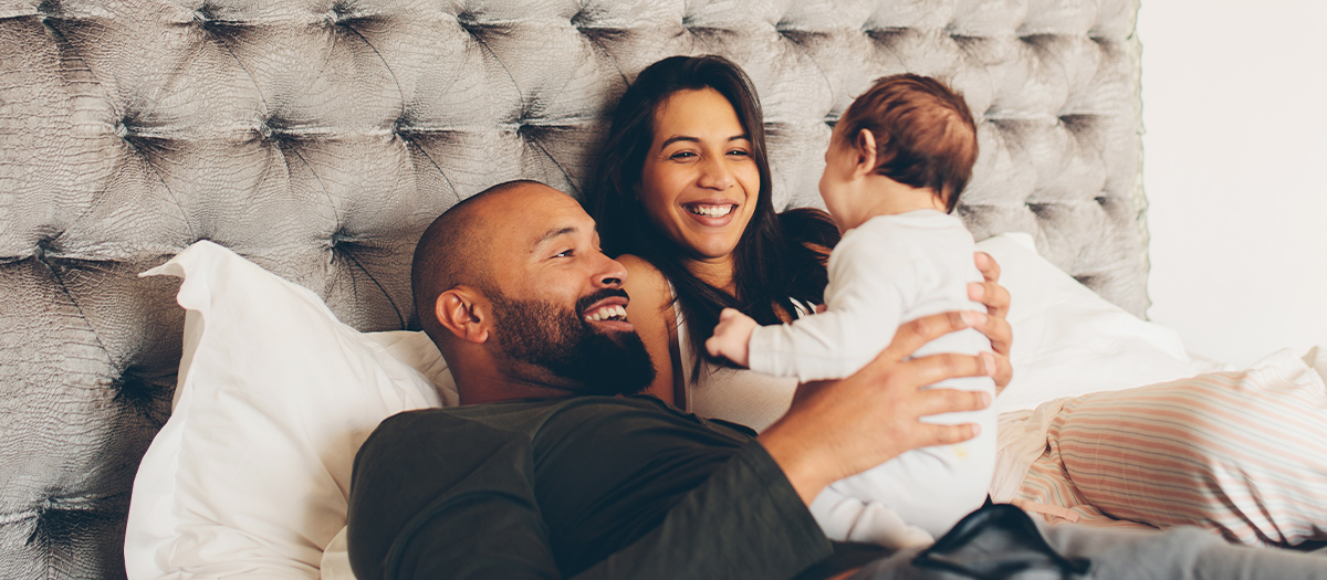 A Mom and dad sitting on bed holding baby up and smiling